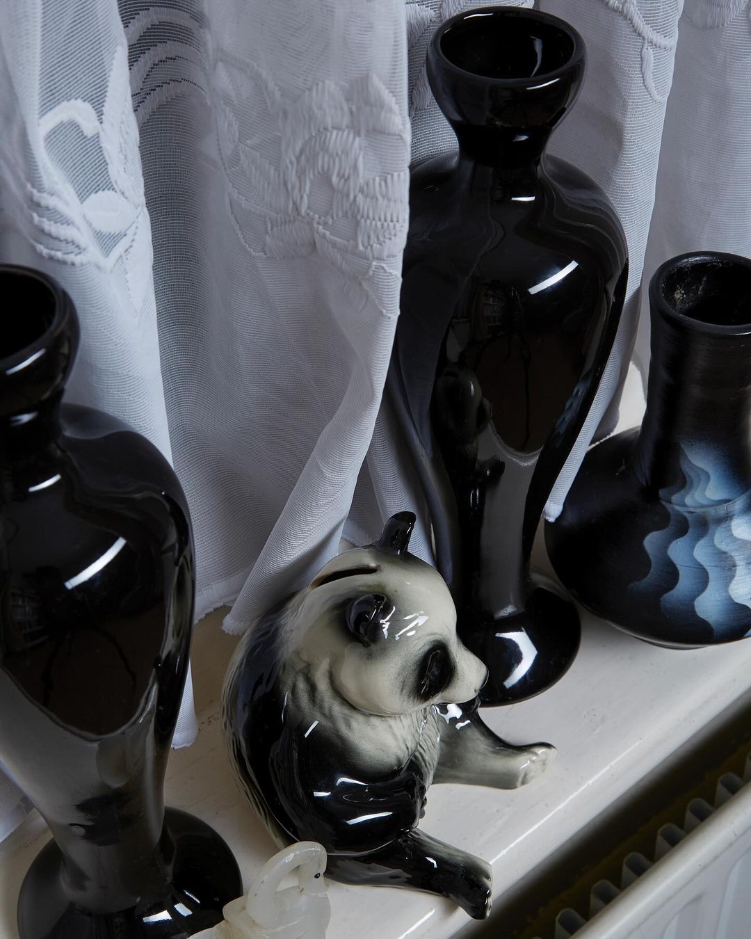 Still life shot features a panda figurine among glossy black vases against a backdrop of semi-sheer white floral patterned curtains.
