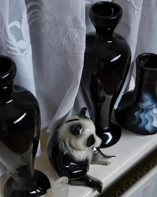 Still life shot features a panda figurine among glossy black vases against a backdrop of semi-sheer white floral patterned curtains.