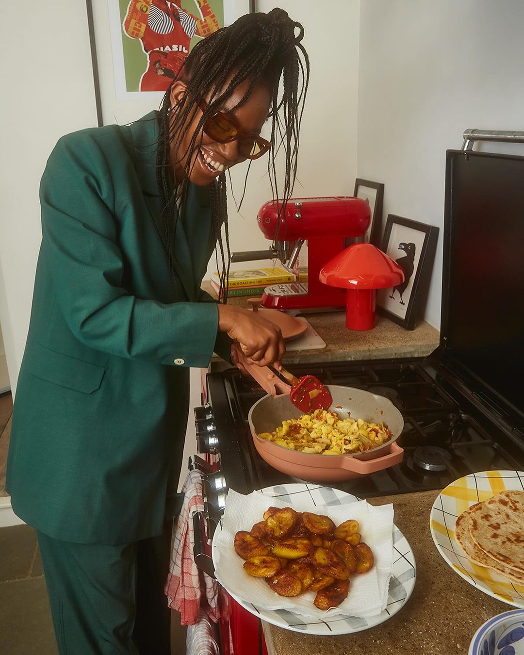 Smiling woman in green suit cooking plantains in a kitchen with red appliances and colorful artwork in the background.