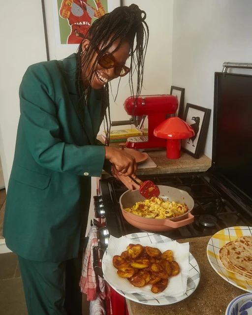 Smiling woman in green suit cooking plantains in a kitchen with red appliances and colorful artwork in the background.