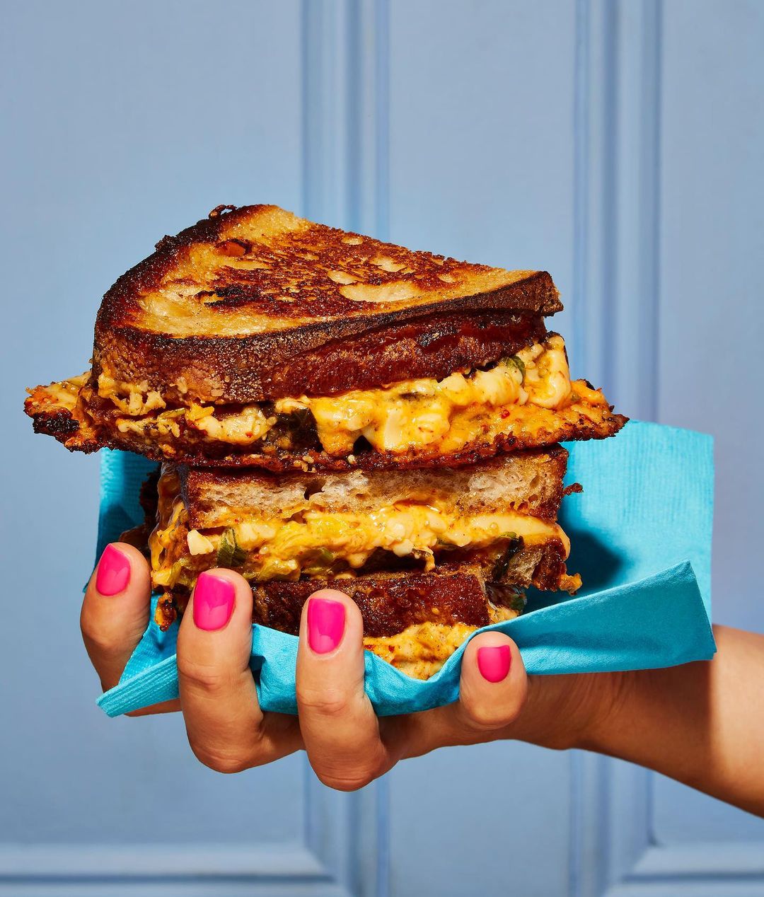 Grilled cheese sandwich held by a hand with pink nail polish against a blue background with paneling in a vertical orientation.