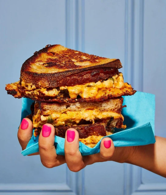 Grilled cheese sandwich held by a hand with pink nail polish against a blue background with paneling in a vertical orientation.
