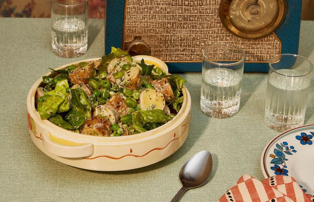 Overhead studio shot of potato salad in a ceramic bowl on a tablecloth, accented by a vintage radio, water glasses, and a spoon.