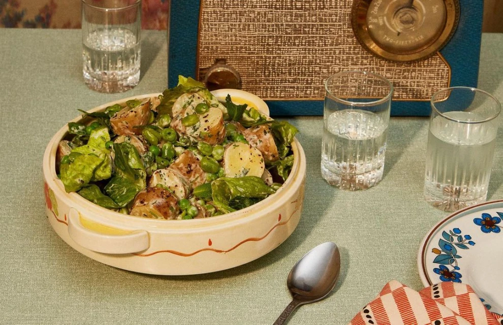 Overhead studio shot of potato salad in a ceramic bowl on a tablecloth, accented by a vintage radio, water glasses, and a spoon.