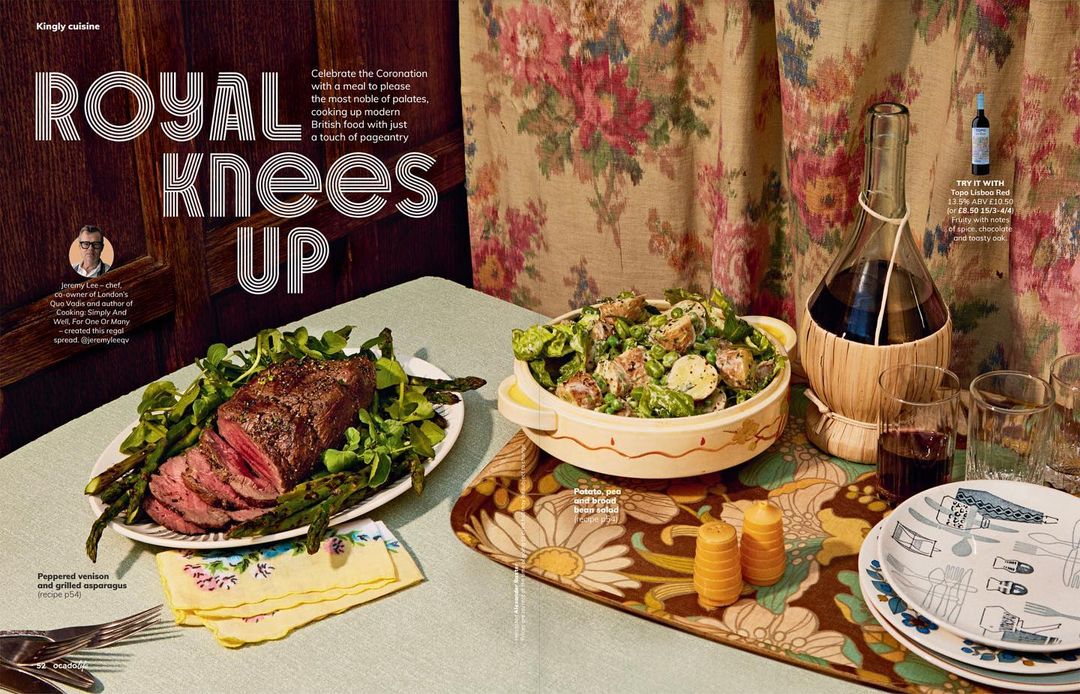 Overhead shot of peppered venison with asparagus, potato salad, and wine on a table with floral wallpaper, creating a refined food display.
