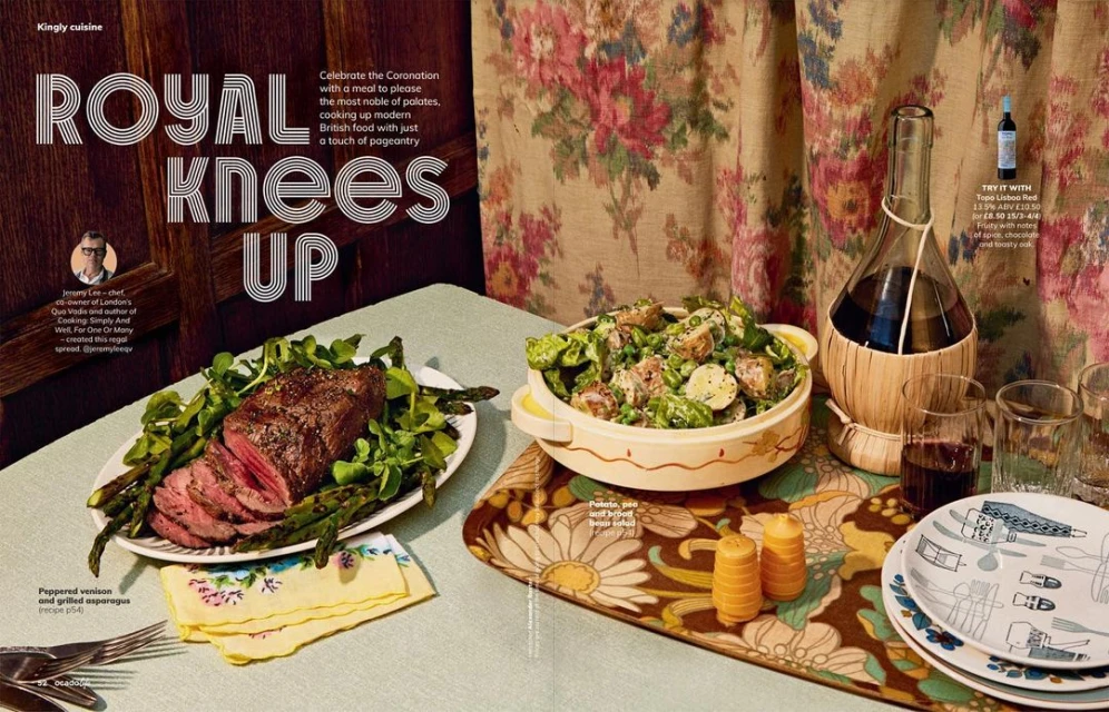 Overhead shot of peppered venison with asparagus, potato salad, and wine on a table with floral wallpaper, creating a refined food display.