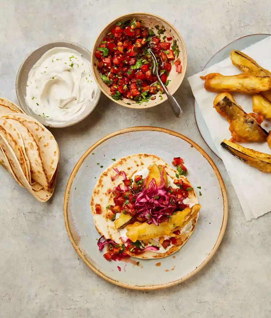 Overhead shot of a fish taco with fried fish, cabbage, tomato salsa, and white sauce on a plate, along with side dishes.