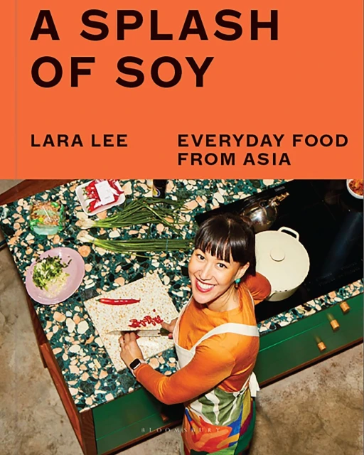 Cookbook cover featuring a smiling woman in an apron at a terrazzo countertop, with vegetables and cooking utensils. Text: A Splash of Soy.