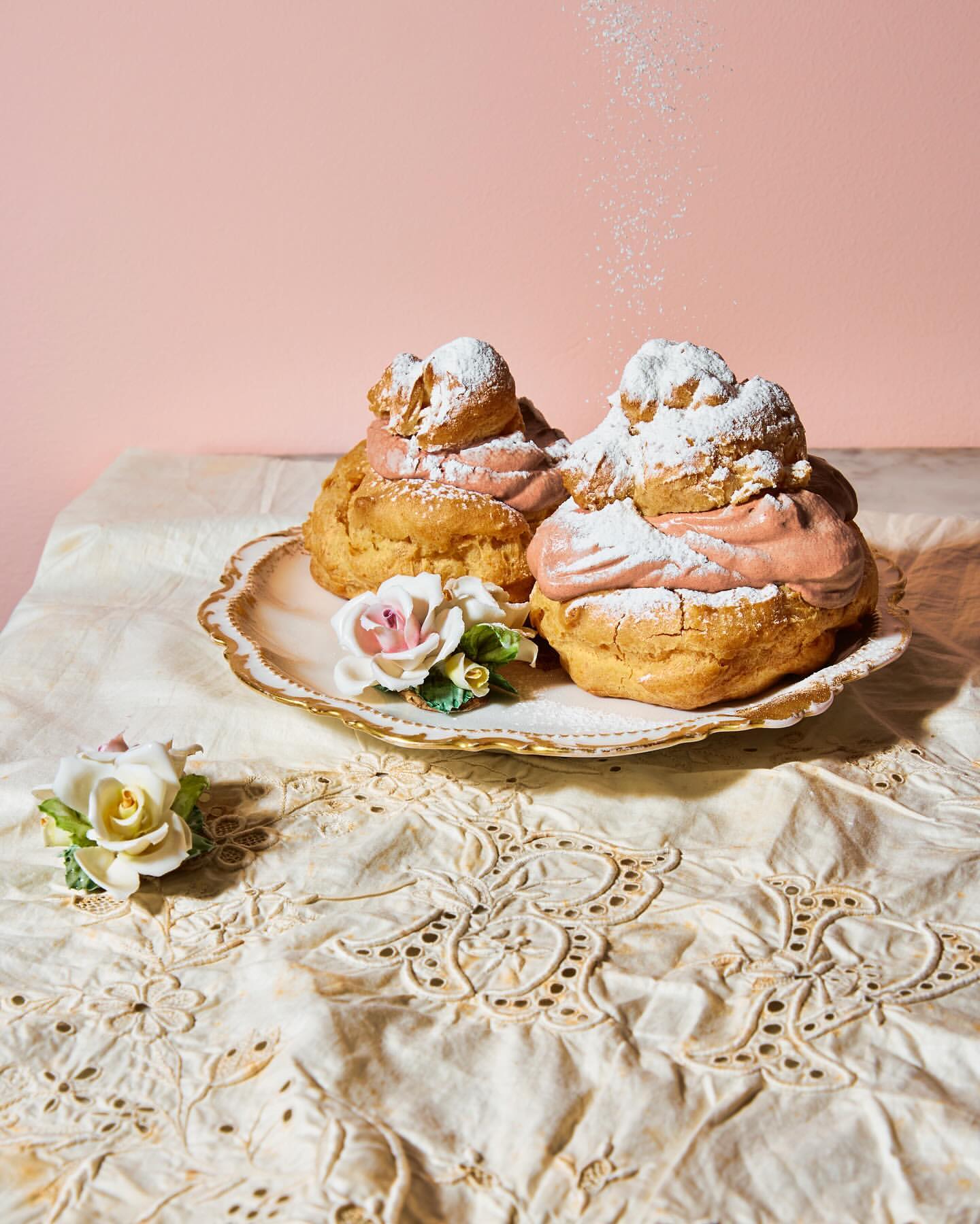 Close-up of two cream puffs dusted with powdered sugar on a decorative plate, accompanied by floral decorations on a lace tablecloth.