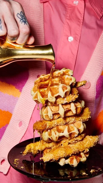 A stack of waffles with syrup being poured on them, held by a person in pink and orange clothing, presenting a visually appealing food scene.