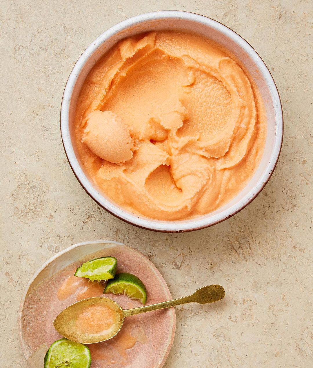 Overhead shot of a bowl of cantaloupe sorbet next to a plate with lime wedges and a spoon on a neutral-colored surface. 