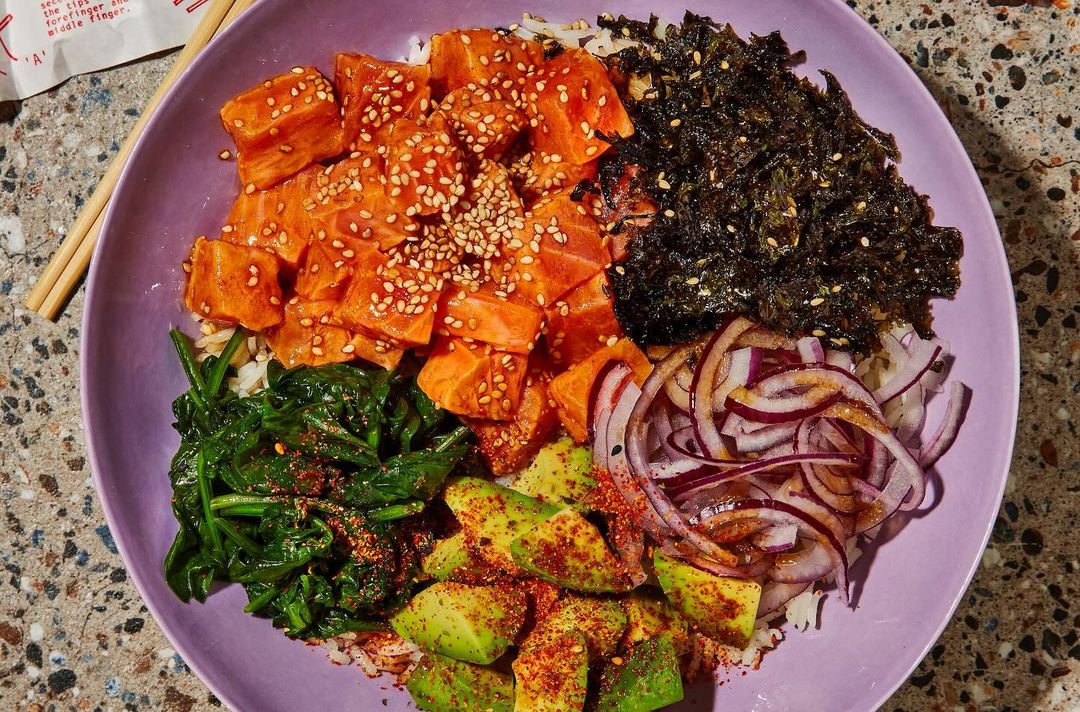 Overhead shot of a vibrant poke bowl with salmon, seaweed, avocado, red onion, and spinach on a lavender plate, and chopsticks.