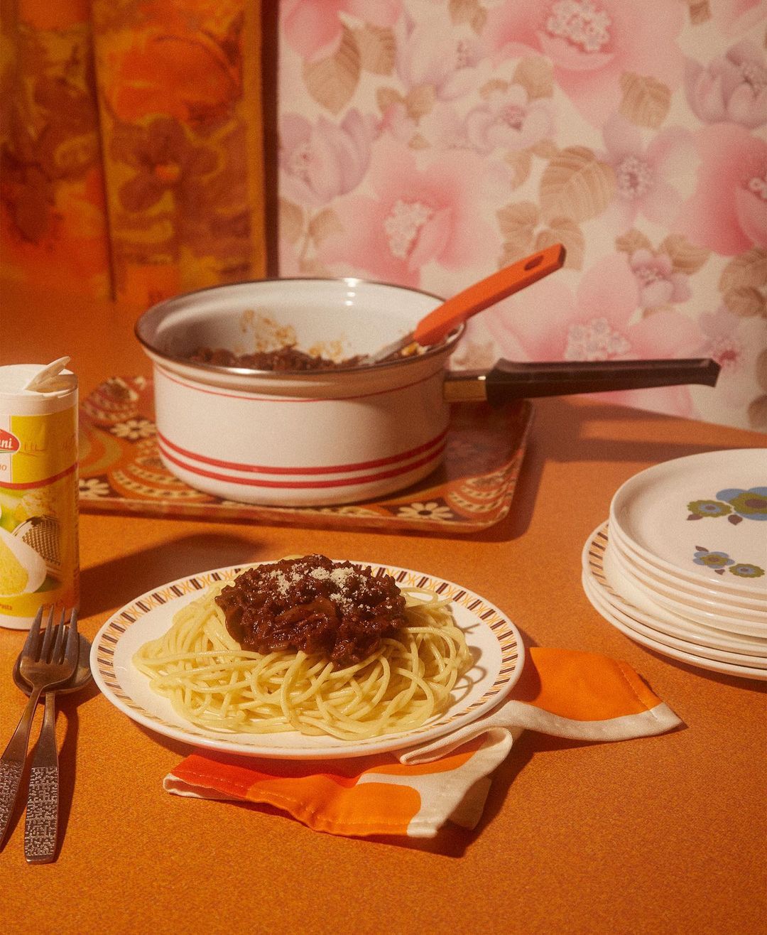 Spaghetti with meat sauce served on a plate on a table with a vintage floral wallpaper background. Cooking pot and utensils nearby.