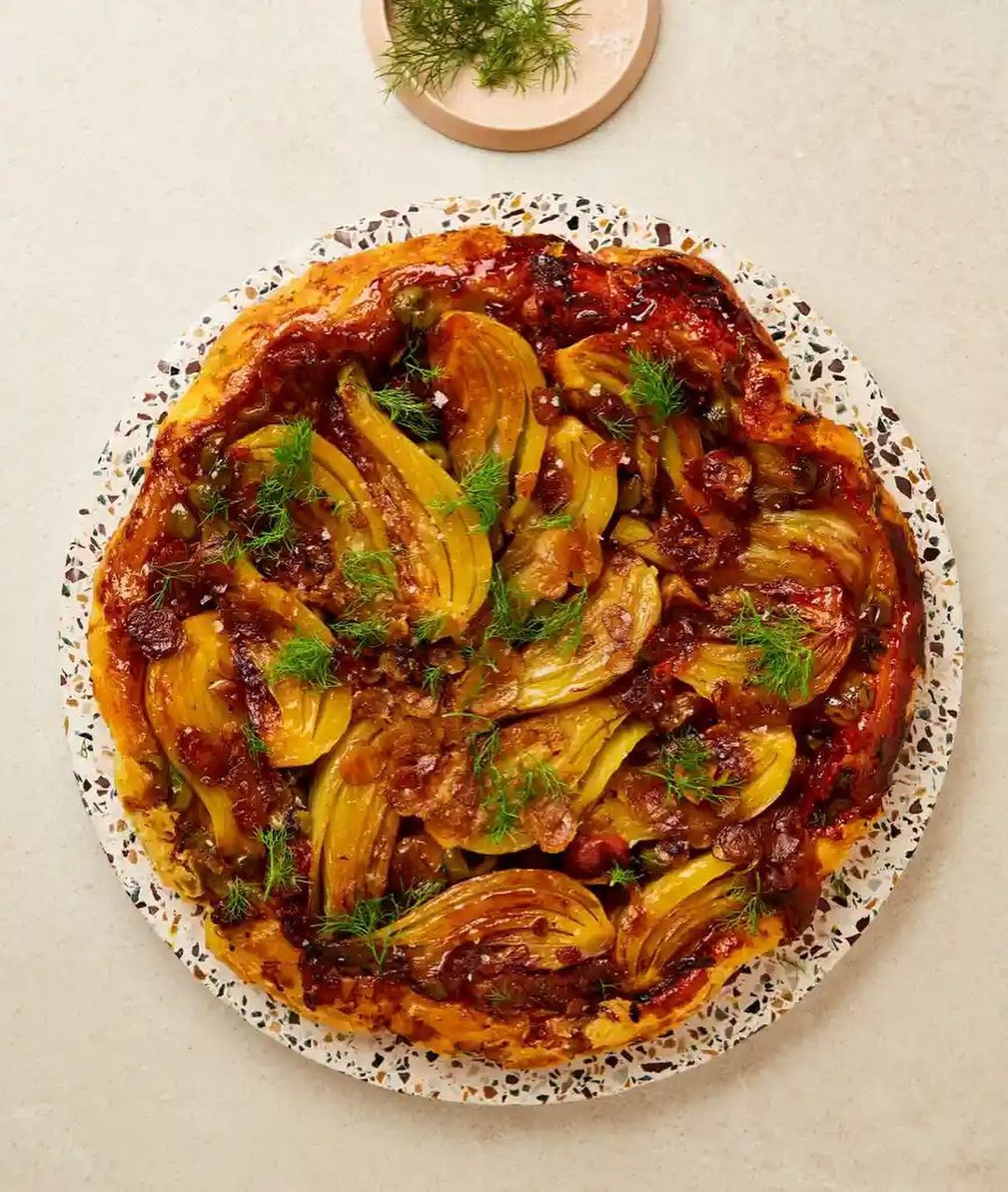 Overhead shot of caramelized fennel tarte tatin on a speckled plate, garnished with fresh dill, set against a neutral backdrop.