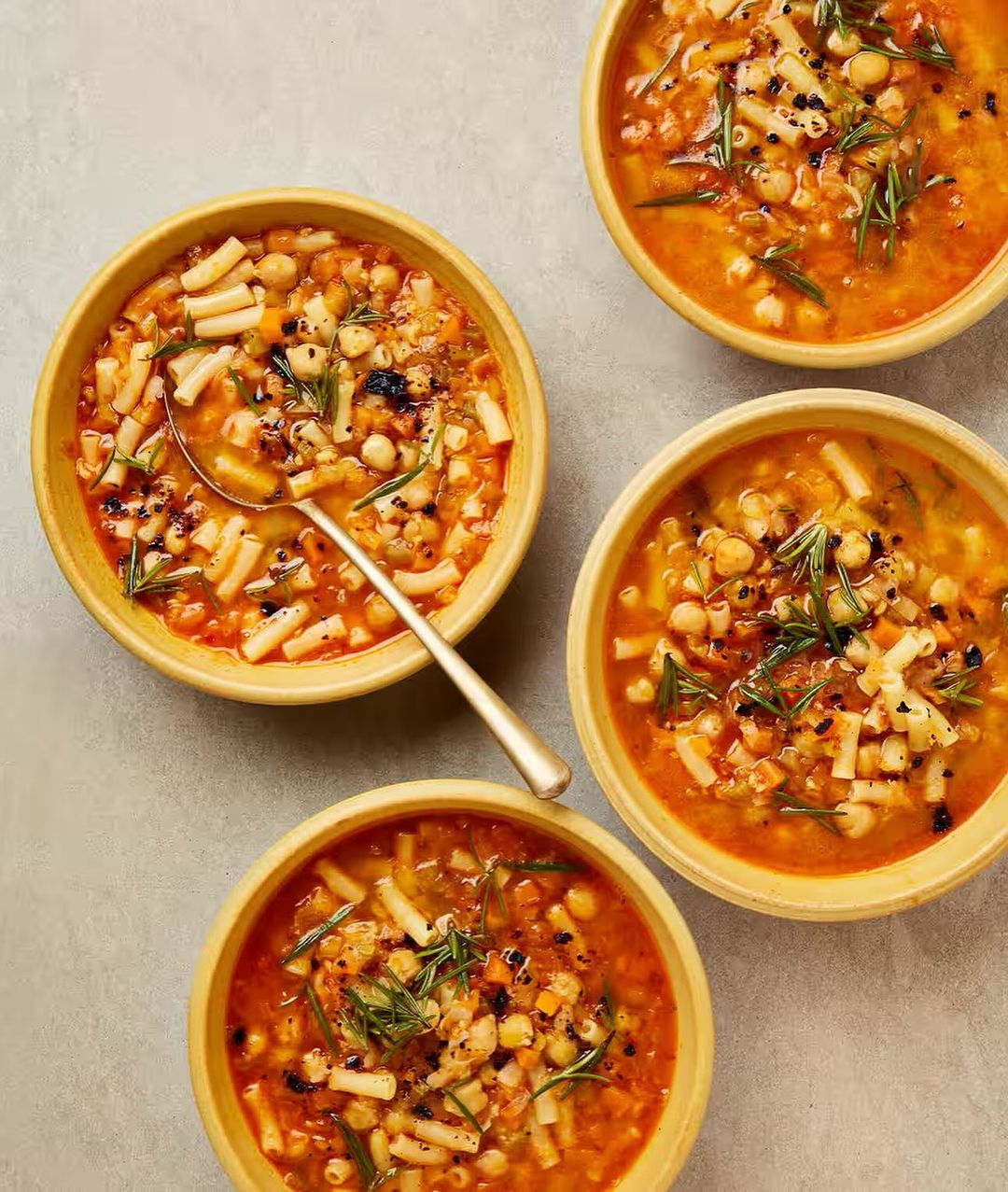 Overhead shot features four bowls of vibrant minestrone soup with pasta, chickpeas, and herbs, highlighting a hearty, comforting meal.