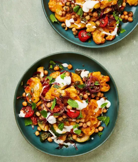 Overhead shot of two teal plates filled with golden cauliflower florets, chickpeas, tomatoes, mint, and a creamy sauce on a muted green table.