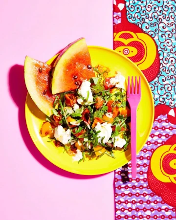 Overhead shot of a vibrant yellow plate featuring a fresh salad with watermelon and mozzarella, alongside a bright pink fork and patterned cloth.