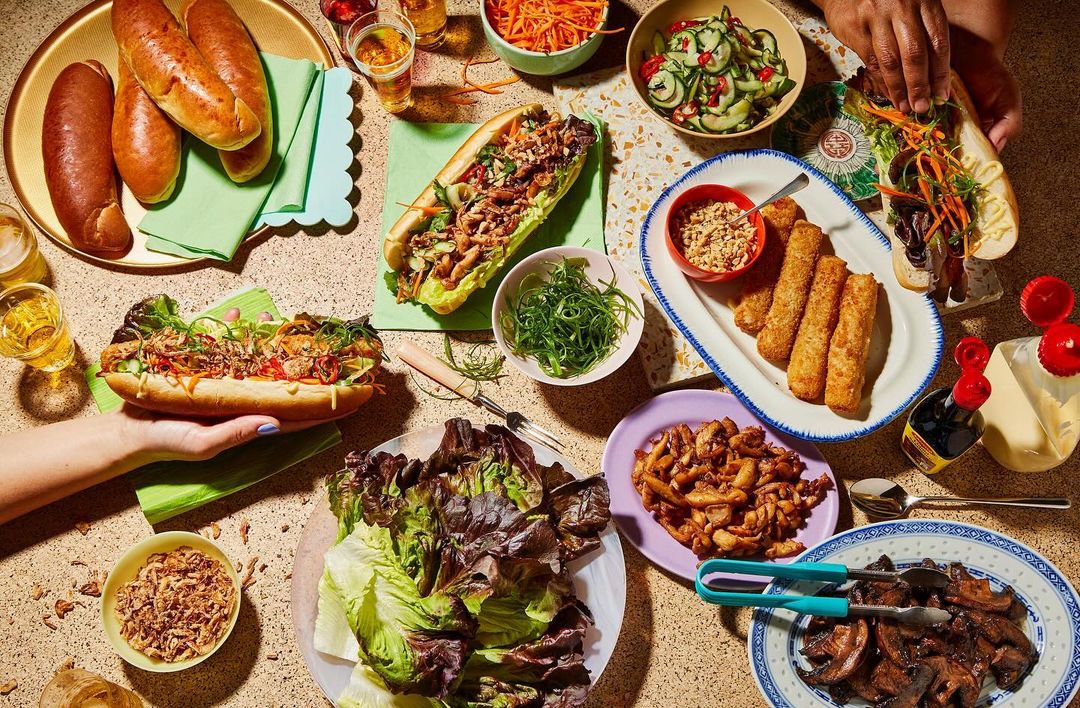 Overhead shot of a table laden with Vietnamese-inspired food: Banh Mi sandwiches, crispy spring rolls, bowls of fresh salads, and assorted beverages.