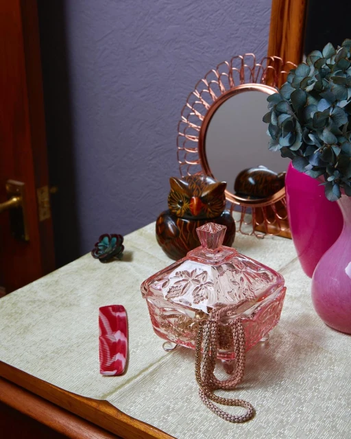 Still life on a table featuring a pink crystal candy dish, an owl figurine, a reflective mirror, and a vase of blue flowers.