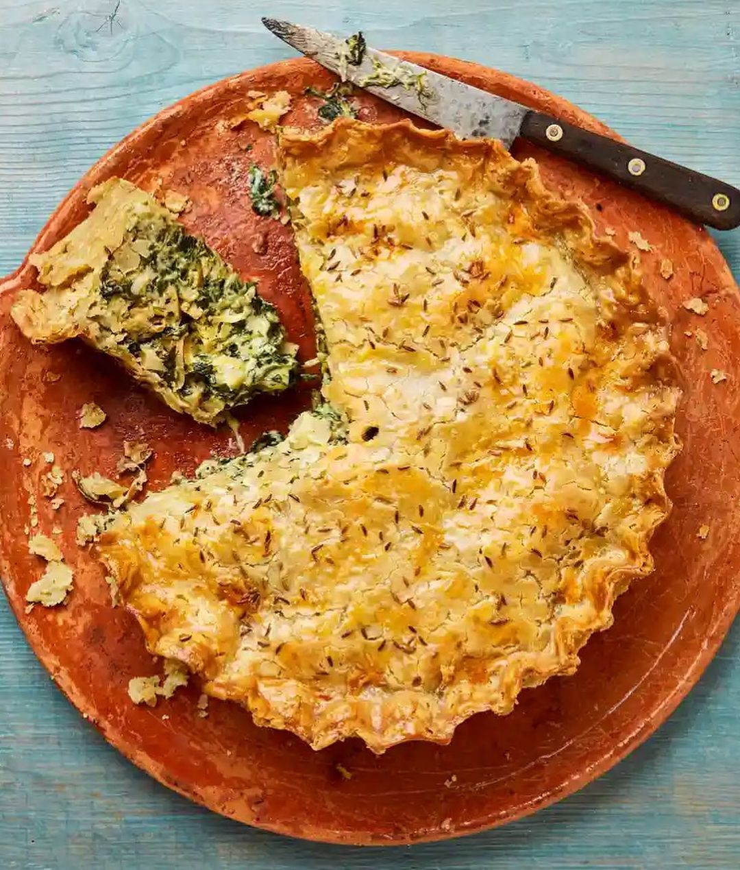 Overhead shot of a partially sliced spinach pie on a terracotta plate with a serving knife on a blue wooden surface.