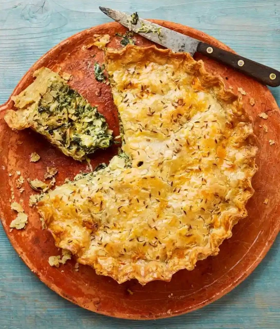 Overhead shot of a partially sliced spinach pie on a terracotta plate with a serving knife on a blue wooden surface.