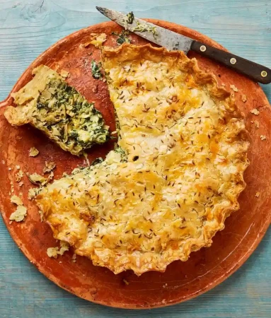 Overhead shot of a partially sliced spinach pie on a terracotta plate with a serving knife on a blue wooden surface.