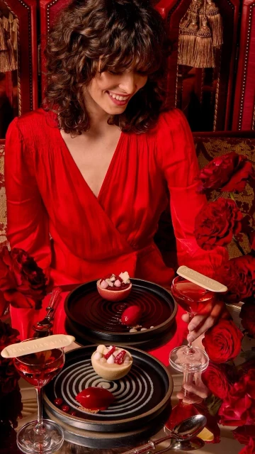 Woman in a red dress smiles while seated at a table with desserts and drinks, surrounded by red roses in a warm, intimate setting.