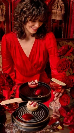 Woman in a red dress smiles while seated at a table with desserts and drinks, surrounded by red roses in a warm, intimate setting.