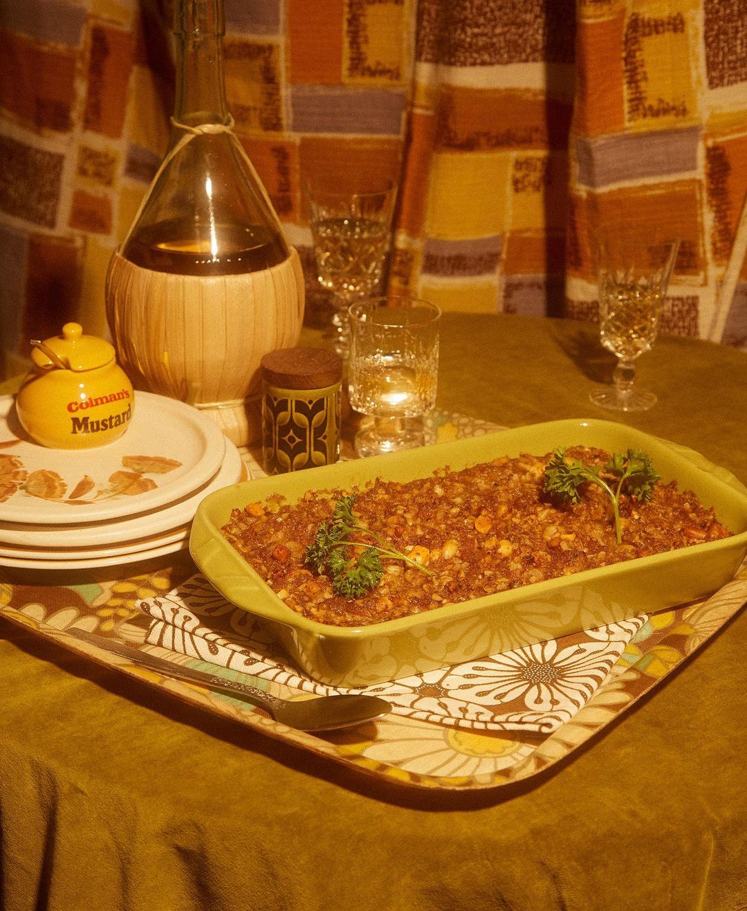 Overhead shot of a casserole dish on a tray with glassware, plates, and condiments set on a dark-gold surface with patterned backdrop.