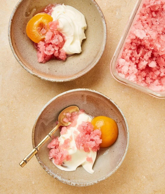 Two bowls of dessert featuring scoops of white cream, granita-style shaved ice, and orange fruit sections, staged on a beige surface.