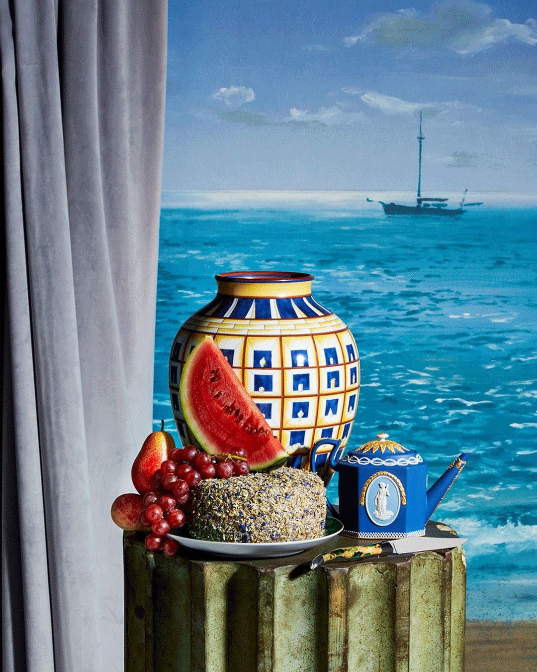 Still life featuring a blue and yellow vase, teapot, cake, fruit, and a slice of watermelon on a column against a sea backdrop.