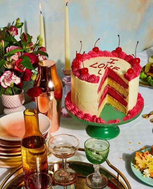 A layer cake on a green cake stand sits among glassware and flowers on a table with tall, lit candles in the background.