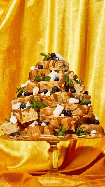 Elevated dessert presentation on a pedestal, showcasing a layered cake composed of stacked square pastry bites with colorful toppings and syrup.