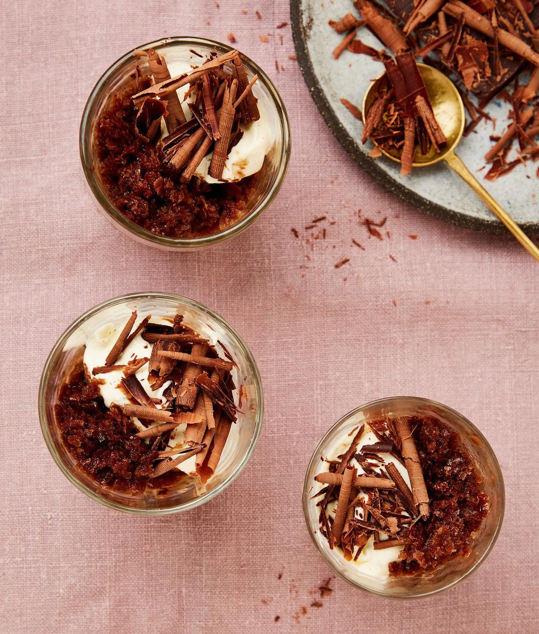 Three glass dessert cups filled with cream, coffee granita and chocolate shavings on a pink tablecloth beside a plate with chocolate.