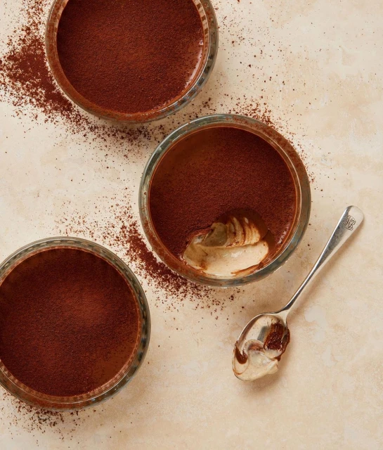 Overhead shot of three glass bowls filled with creamy dessert topped with cocoa powder, accompanied by a silver spoon on a beige surface.