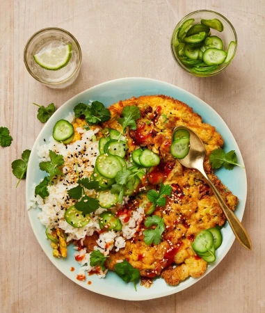 Overhead shot of a vibrant egg foo young dish with rice, cucumbers, and cilantro served on a blue plate, complemented by drinks.