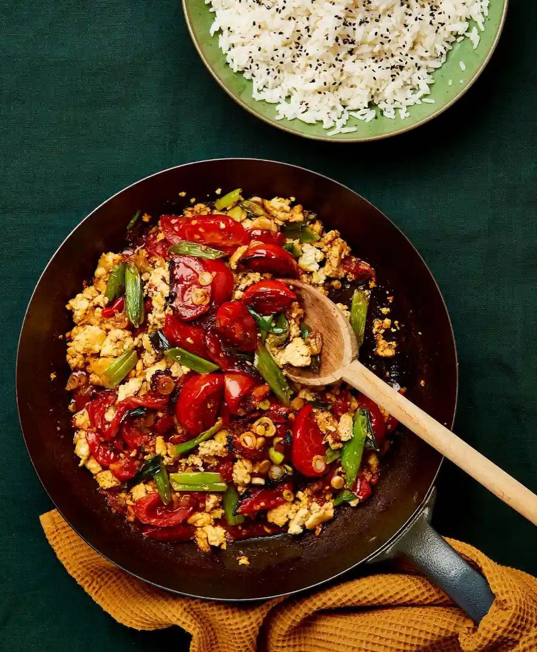 Overhead shot of stir-fried tomatoes, tofu, and scallions in a wok, with a wooden spoon and a side of rice on a dark green tablecloth.