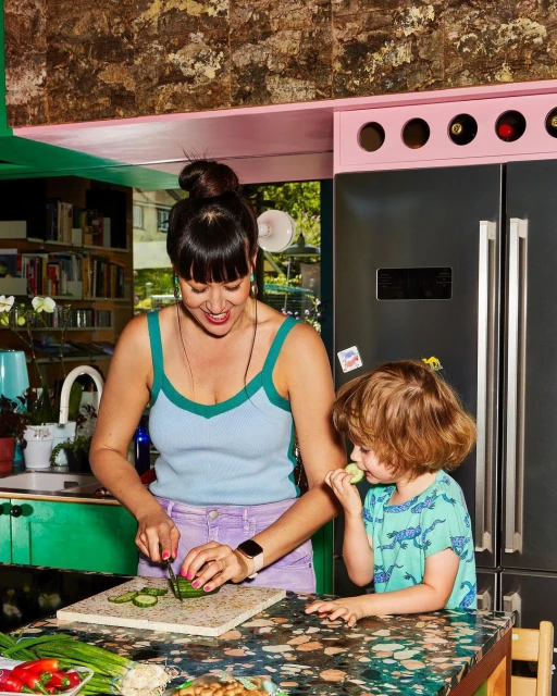 Woman cuts cucumber at kitchen counter as a child eats cucumber beside her, with a refrigerator and shelving visible in the background.