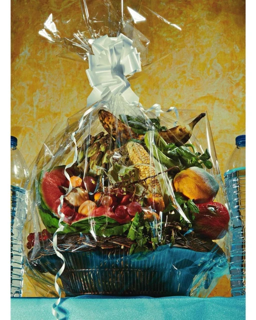 Gift basket filled with fruit and vegetables, wrapped in cellophane with a white bow on top, set against a textured yellow backdrop.