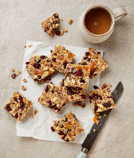 Overhead view of granola bars with dried fruit and nuts served on parchment paper with a knife and a mug of coffee.