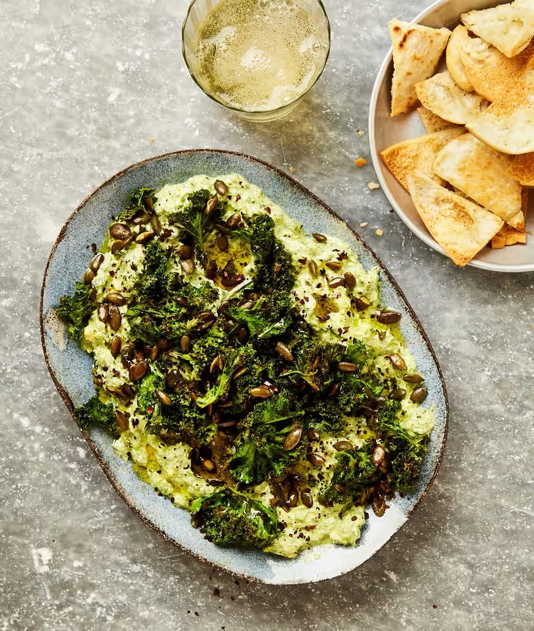 Overhead shot of a blue plate filled with green dip, kale, and seeds, along with a glass of drink and bowl of chips.