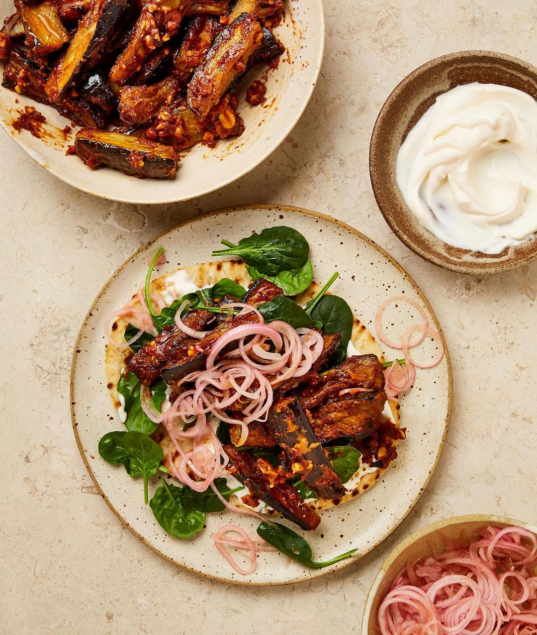 Overhead shot of flatbread topped with eggplant, spinach, and red onion rings, next to bowls of eggplant and yogurt sauce.