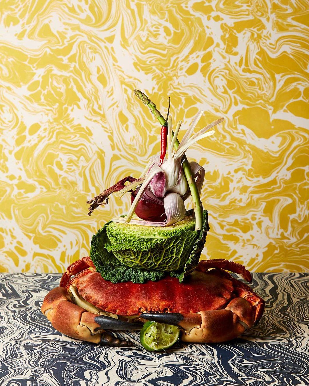 Still life featuring a crab topped with vegetables like cabbage, garlic, and chili peppers on a patterned surface with patterned wallpaper.