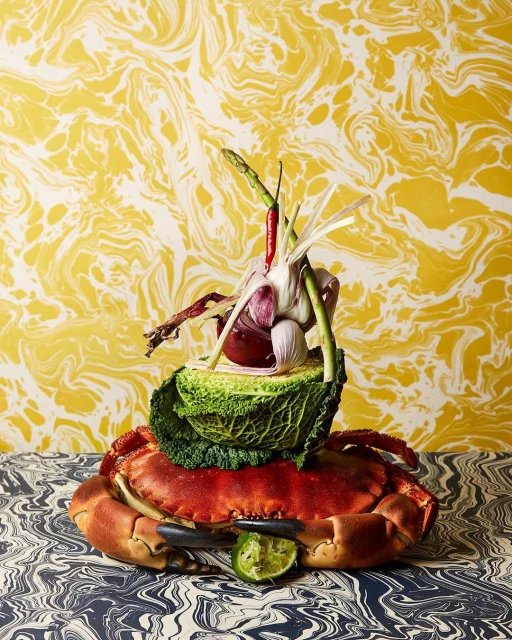 Still life featuring a crab topped with vegetables like cabbage, garlic, and chili peppers on a patterned surface with patterned wallpaper.