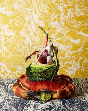 Still life featuring a crab topped with vegetables like cabbage, garlic, and chili peppers on a patterned surface with patterned wallpaper.