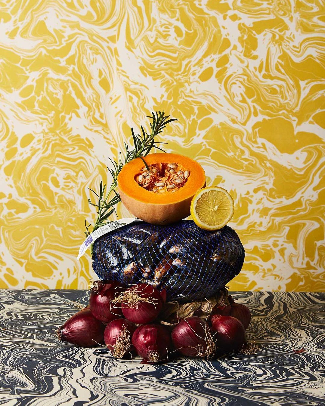 Still life featuring a pumpkin half, lemon, and red onions piled high, set against a marbled yellow and white background.