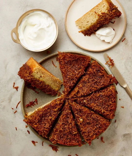 Overhead shot of a coconut cake cut into slices on a plate, with a knife and bowl of white cream on a neutral surface.