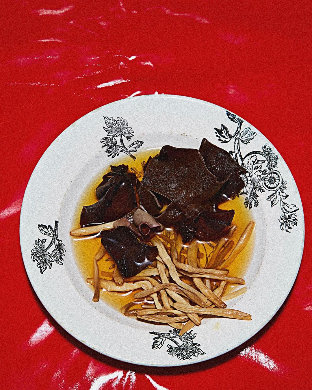 Overhead shot of white plate with patterned rim holding dark mushrooms and light sprouts in broth, set against glossy red background.