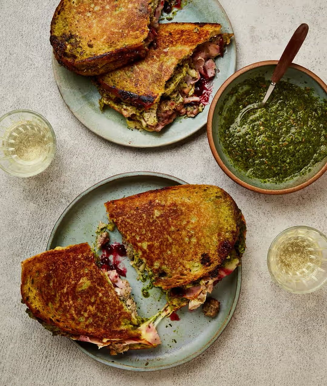 Overhead shot of sliced grilled cheese sandwiches filled with meat and sauce on gray plates with a bowl of pesto and drinks.