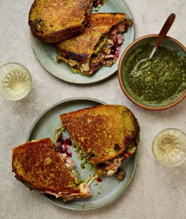 Overhead shot of sliced grilled cheese sandwiches filled with meat and sauce on gray plates with a bowl of pesto and drinks.